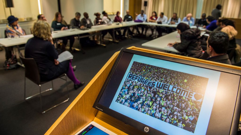 Close up of computer screen with image saying refugees welcome. In background a classroom with students sitting around the table